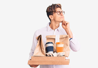 Young handsome man wearing business clothes holding delivery food serious face thinking about question with hand on chin, thoughtful about confusing idea