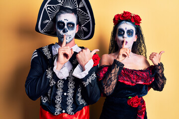 Young couple wearing mexican day of the dead costume over yellow asking to be quiet with finger on lips pointing with hand to the side. silence and secret concept.