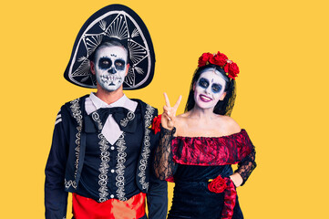 Young couple wearing mexican day of the dead costume over background showing and pointing up with fingers number two while smiling confident and happy.