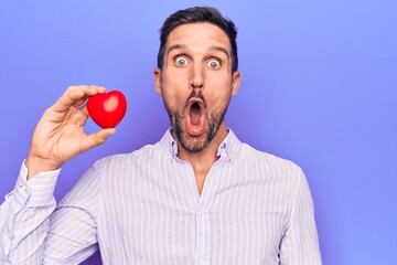 Young handsome man asking for health care holding red heart over isolated purple background scared and amazed with open mouth for surprise, disbelief face