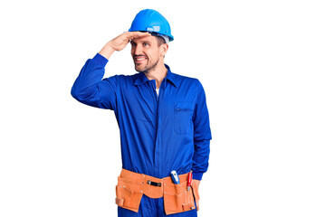 Young handsome man wearing worker uniform and hardhat very happy and smiling looking far away with hand over head. searching concept.