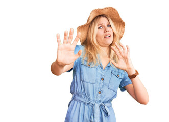 Young beautiful blonde woman wearing summer hat and dress afraid and terrified with fear expression stop gesture with hands, shouting in shock. panic concept.