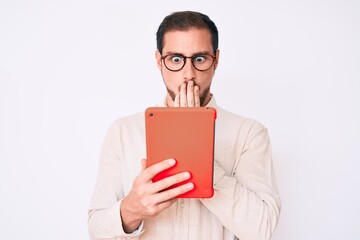 Young handsome man using touchpad device covering mouth with hand, shocked and afraid for mistake....