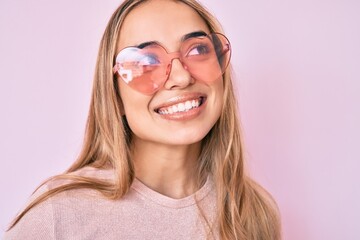 Young beautiful blonde woman wearing heart shaped sunglasses smiling looking to the side and staring away thinking.