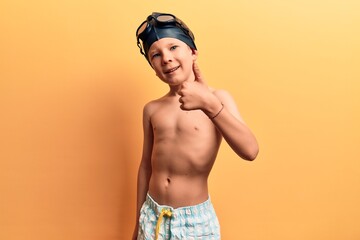 Cute blond kid wearing swimwear and swimmer glasses smiling happy and positive, thumb up doing excellent and approval sign