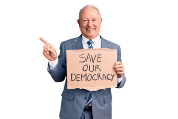 Senior handsome grey-haired man holding save our democracy cardboard banner smiling happy pointing with hand and finger to the side
