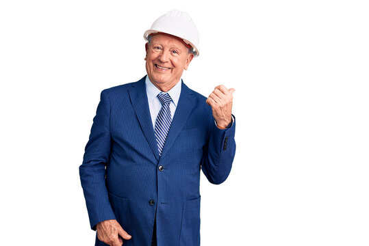 Senior handsome grey-haired man wearing suit and architect hardhat smiling with happy face looking and pointing to the side with thumb up.