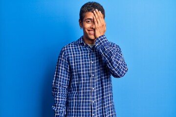 Young african amercian man wearing casual clothes covering one eye with hand, confident smile on face and surprise emotion.