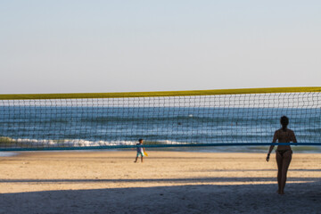 Rede de volley na praia em primeiro plano com mar ao fundo e criança desfocada e silhueta e sombra de uma mulher.