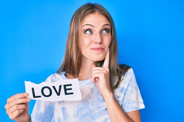 Young blonde girl holding paper with love message serious face thinking about question with hand on...