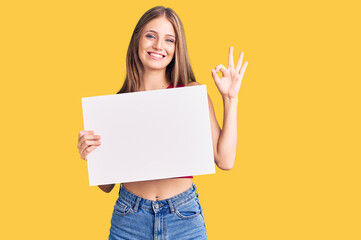 Obraz na płótnie Canvas Young beautiful blonde woman holding blank empty banner doing ok sign with fingers, smiling friendly gesturing excellent symbol