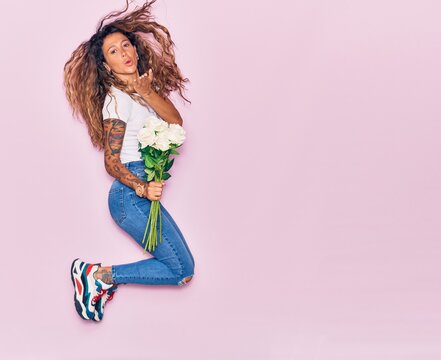 Young beautiful curly woman with tattoo holding bouquet of white rose smiling happy. Jumping with smile on face throwing kiss with hand over isolated pink background