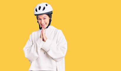 Beautiful brunette young woman wearing bike helmet and sporty clothes praying with hands together asking for forgiveness smiling confident.