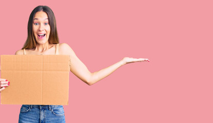 Beautiful brunette young woman holding cardboard banner with blank space celebrating victory with happy smile and winner expression with raised hands