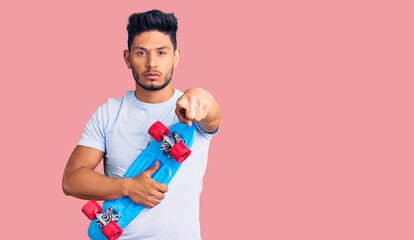 Handsome latin american young man holding skate pointing with finger to the camera and to you, confident gesture looking serious