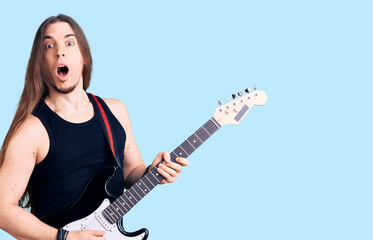 Obraz premium Young adult man with long hair playing electric guitar scared and amazed with open mouth for surprise, disbelief face