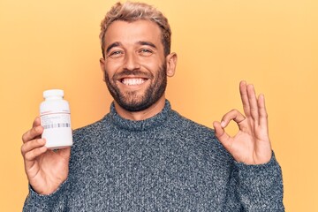 Young handsome blond man with beard holding jar of medicine pills over yellow background doing ok sign with fingers, smiling friendly gesturing excellent symbol