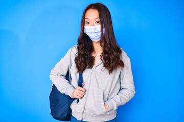 Young beautiful chinese girl holding student backpack wearing medical mask scared and amazed with open mouth for surprise, disbelief face