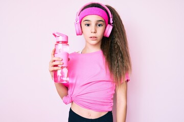 Cute hispanic child girl wearing gym clothes and using headphones holding bottle of water thinking attitude and sober expression looking self confident