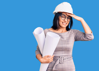 Young beautiful latin woman wearing architect hardhat and glasses holding blueprints stressed and frustrated with hand on head, surprised and angry face