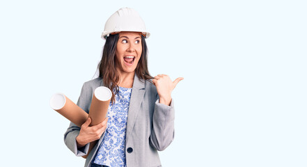Young beautiful brunette woman wearing architect hardhat holding blueprint pointing thumb up to the side smiling happy with open mouth