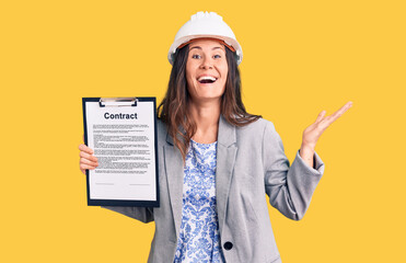 Young beautiful brunette woman wearing architect hardhat holding contract celebrating victory with happy smile and winner expression with raised hands