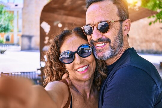 Middle age beautiful couple wearing casual clothes and sunglasses smiling happy. Standing with smile on face making selfie by the camera hugging at town street.