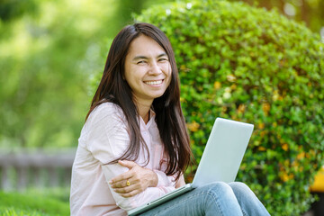 Asian woman sitting green park using laptop computer. Woman working on laptop happy entrepreneur business using notebook with hands typing on keyboard home office during coronavirus quarantine period