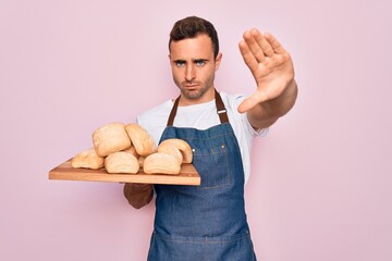 Young handsome baker man with blue eyes wearing apron holding tray with homemade bread with open hand doing stop sign with serious and confident expression, defense gesture