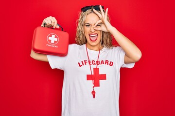 Beautiful blonde lifeguard woman wearing t-shirt with red cross and whistle holding first aid kit...