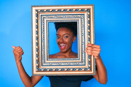 Young african american woman holding empty frame pointing thumb up to the side smiling happy with open mouth