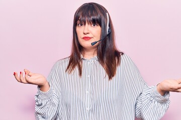 Young plus size woman wearing call center agent headset clueless and confused expression with arms...