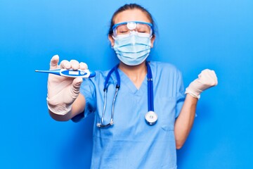 Young blonde woman wearing doctor uniform and coronavirus protection holding help reminder screaming proud, celebrating victory and success very excited with raised arm