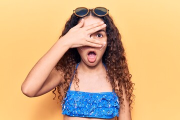 Beautiful kid girl with curly hair wearing bikini and sunglasses peeking in shock covering face and eyes with hand, looking through fingers afraid