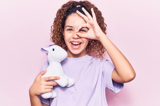Beautiful kid girl with curly hair holding animal doll toy smiling happy doing ok sign with hand on eye looking through fingers