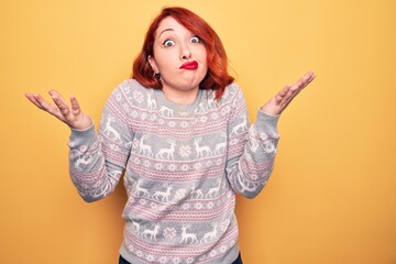 Beautiful redhead woman wearing christmas sweater with reindeer over yellow background clueless and...
