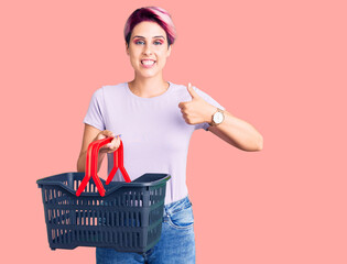 Young beautiful woman with pink hair holding supermarket shopping basket smiling happy and positive, thumb up doing excellent and approval sign