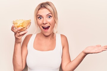 Young beautiful blonde woman holding bowl with potatoes chips over isolated white background celebrating achievement with happy smile and winner expression with raised hand