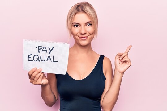 Beautiful blonde woman asking for equality economy holding paper with pay equal message smiling happy pointing with hand and finger to the side