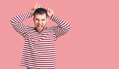 Young handsome man wearing striped sweater posing funny and crazy with fingers on head as bunny ears, smiling cheerful