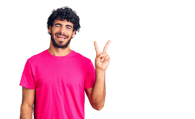 Handsome young man with curly hair and bear wearing casual pink tshirt smiling with happy face winking at the camera doing victory sign. number two.