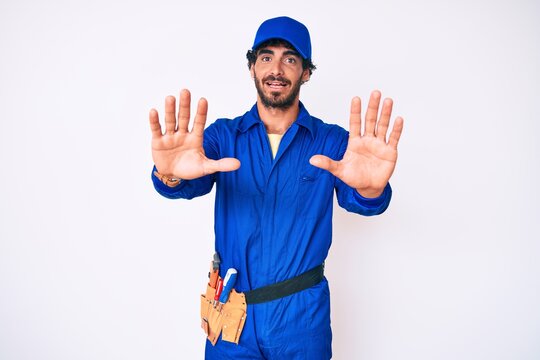 Handsome young man with curly hair and bear weaing handyman uniform doing stop gesture with hands palms, angry and frustration expression