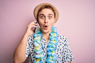 Young man on vacation wearing hat and hawaiian lei having conversation talking on smartphone scared...
