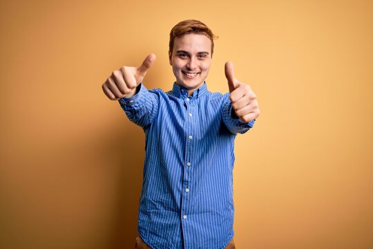 Young handsome redhead man wearing casual striped shirt over isolated yellow background approving doing positive gesture with hand, thumbs up smiling and happy for success. Winner gesture.