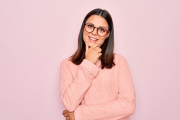 Young beautiful brunette woman wearing casual sweater and glasses over pink background looking confident at the camera smiling with crossed arms and hand raised on chin. Thinking positive.