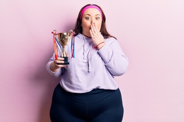 Young plus size woman holding winner trophy covering mouth with hand, shocked and afraid for mistake. surprised expression