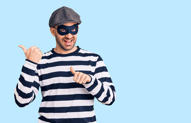 Fototapeta premium Young handsome man wearing burglar mask pointing to the back behind with hand and thumbs up, smiling confident