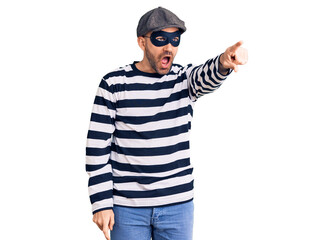 Young handsome man wearing burglar mask pointing with finger surprised ahead, open mouth amazed expression, something on the front