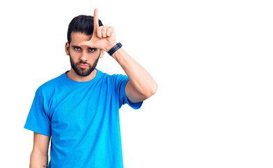 Young handsome man with beard wearing casual t-shirt making fun of people with fingers on forehead doing loser gesture mocking and insulting.
