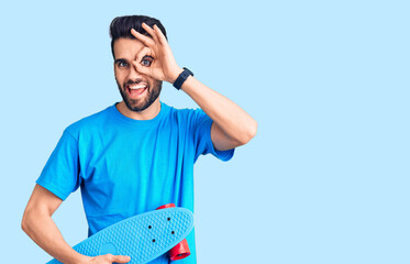 Young handsome man with beard holding skate smiling happy doing ok sign with hand on eye looking through fingers